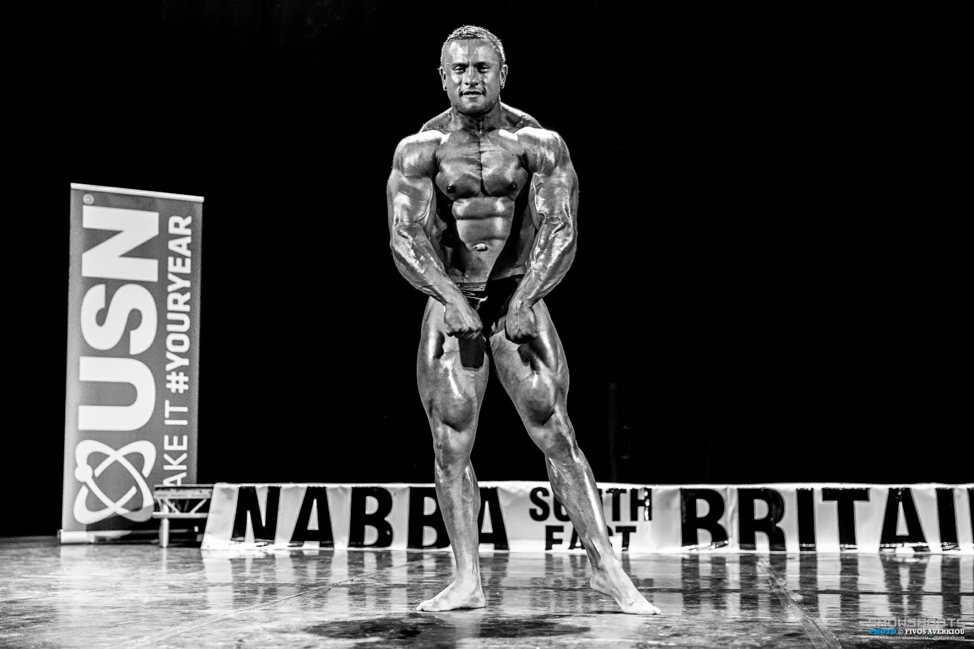 Domenico Monk performing a front pose on the NABBA South Britain stage in black and white.