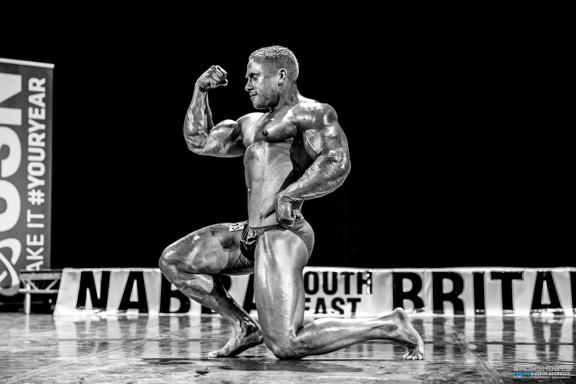 Black and white competition shot — kneeling one-arm bicep flex under stage light